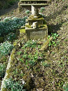 Un marciapiede fatiscente circonda una tomba in stile con una tavoletta e una lapide a croce caduta.