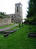 The east end of a stone church seen through a graveyard from the north. On the right is a battlemented tower and the nave extends to the left