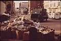 SANITATION DEPARTMENT TRUCK PREPARES TO HAUL ACCUMULATED TRASH FROM STREET. BANNER IN BACKGROUND REMINDS CITIZENS... - NARA - 545502.jpg