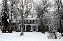 A snow-covered 2-story house with dormer windows on the roof and a porch on the ground floor. The stone house has several large trees in front.