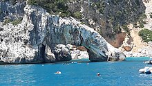 Natural white and grey rocks form a large arch among a bright blue sea with swimmers. The arch and the beige rock behind are covered in scrub.