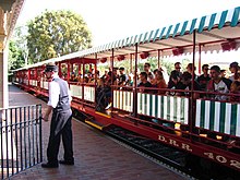 A series of red-colored gondola train cars with green-and-white-striped awnings. A train conductor is in the foreground.