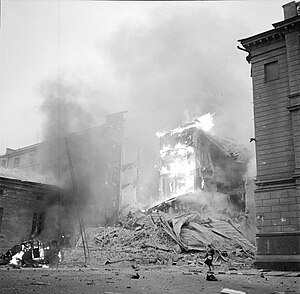 An apartment building is on fire and has partly collapsed in central Helsinki after the Soviet aerial bombing on 30 November 1939. A woman in a coat and a hat is passing on the right next to the rubble, and a car is on fire on the left.