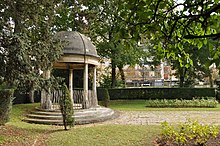 Der Tempel der Liebe von links gesehen, im Schatten befindet er sich auf der linken Seite.  Rechts ein kleiner Teil des Art-Deco-Gartens.  Im Hintergrund hohe Bäume und Gebäude an der Avenue de Madrid.