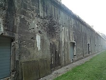 Uma foto de uma parte da vala do desfiladeiro do Fort de Loncin.  Há uma cratera em forma de concha na parede no centro superior da imagem.