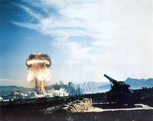 An artillery piece with a mushroom cloud rising in the distance