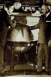 Smith (usando óculos) e Truman se inclinam sobre um grande globo.  Um relógio no consolo da lareira atrás do globo indica que são dez horas.