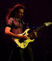 A man wearing black clothing and a chain necklace, holding an electric guitar