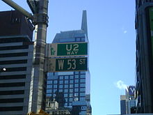 A green street sign with 53rd Street written on it. Just above the sign is another, identical in colour, which says "U2 Way". A skyscraper with reflective windows is immediately behind the signs.
