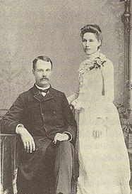 Photographie en noir et blanc d'un couple.  À gauche, un homme assis avec une moustache portant un costume sombre.  Debout à droite, une femme en robe blanche, le corps tourné vers la gauche.  Les deux font face à la caméra.