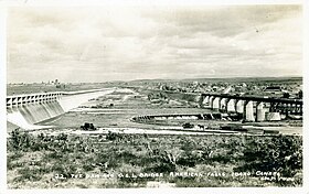 View across a river, with a concrete dam on the left and a bridge on the right with a town on the opposite bank.