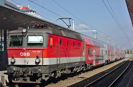 1144 230 vor einem Doppelstockzug fährt als R 2325 Richtung Payerbach-Reichenau am Bahnsteig 2 des Bahnhofs Wien Praterstern ein