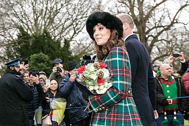 A woman, who is smiling slightly and holding a bouquet of flowers, standing in the foreground, with a police officer and a crowd of people taking photographs in the background