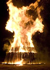 De grandes flammes engloutissent la couche supérieure d'une structure circulaire à trois niveaux de bûches;  les rondins sont à la verticale du sol.