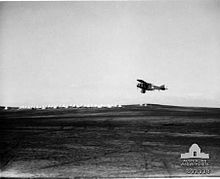 Aircraft flying over flat ground with tents and buildings on skyline