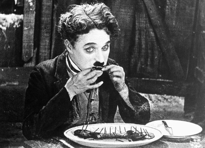 Chaplin, assis à table, mangeant un bout de sa chaussure.