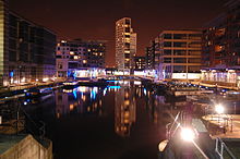 A night-time scene of a dock containing a number of moored canal-boats to left and right and railings around the edges. At the front is part of a lock gate and steps leading down to the water. Around most of the dock are multi-storey modern buildings, some with lighted ground-floors and seats and decorative objects outside. The most prominent of these, at the far end, is a twenty-storey building with curving facades.