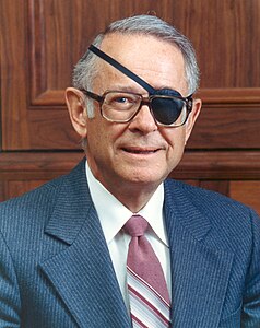 Photo en couleur d'un homme portant veston. Son oeil gauche est recouvert d'un bandeau et il porte des lunettes.