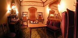 Petit salon dans lequel se trouve une table au centre, une chaise et une cheminée sur la gauche, une porte entourée de deux meubles au fond et, sur la droite, un sofa surmonté d'un miroir ainsi qu'un paravent, le tout dans le style art nouveau