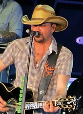 A young man in a checked shirt and a cowboy hat playing a guitar and singing into a microphone