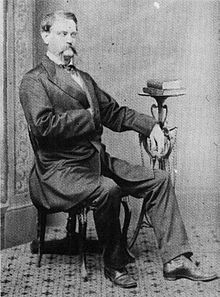 A mustachioed man sitting with his right hand in his waistcoat and his left arm resting on a bookstand