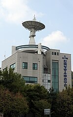Zdjęcie budynku z dużą anteną na dachu. Budynek z szarych płytek z dużymi oknami i niebieskim, koreańskim napisem.