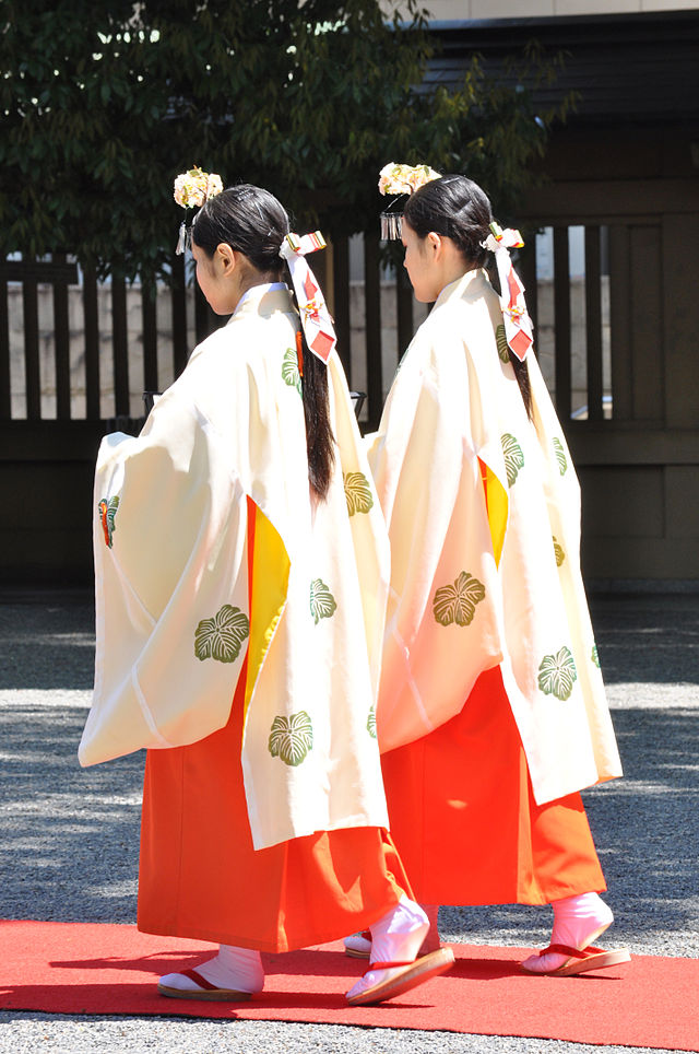 Photo couleur de deux jeunes femmes de trois-quart dos, debout, portant un haut blanc, une sorte de jupe longue rouge, et une épingle à cheveux ornée de fleurs au-dessus de leur front.