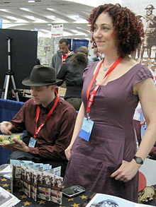 Una donna con i capelli rossi ricci in piedi dietro una scrivania che saluta le persone