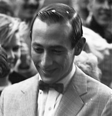 A smiling man looking down with slicked back hair. He is wearing a suit with a bow tie.