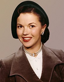 Young, smiling, dark-haired woman wearing a hat and business attire, with a double strand of pearls around her neck