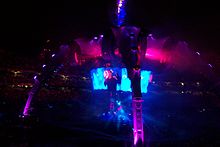 At night in a large outdoor stadium filled with people, a structure with four legs curving upward and meeting in a central spire is bathed in indigo and violet light, as a large circular video screen below the spire but above a stage shows a dark-clothed man with arm raised singing into an illuminated circular microphone.