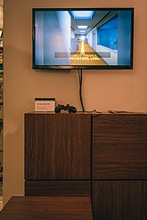 Televizní obrazovka umístěná nad transparentem s ovladačem vedle něj.  Televize zobrazuje část „Konec zmatků“ remaku, ve kterém je hráči řečeno, aby sledoval malovanou žlutou čáru přes kancelář.