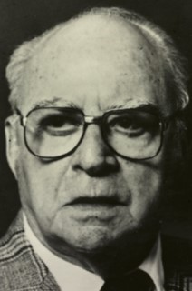 Black and white photo of a middle-aged man, balding with grey hair; wearing dark-rimmed eyeglasses, a checkered suitjacket, a white dress shirt and a dark-coloured neck tie.