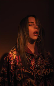 A photograph of a black-haired Eilish wearing a red top in front of a dark backdrop.