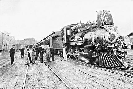 "Seattle, Lake Shore and Eastern Railway's Engine No. 2, the D.H. Gilman, photographed on Independence Day, 1895", "despite the rain", at Columbia Street Station on Railroad Avenue built on pilings over filled mudflat, now Western Avenue. The occasion had a holiday excursion to Sumas. The quote is from the foreground of the image.