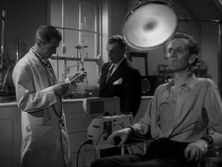 Quatermass (Brian Donlevy) (centre) and Briscoe (David King-Wood) (left) examine Carroon (Richard Wordsworth) (seated) in a scene from The Quatermass Xperiment. Makeup artist Phil Leakey worked with cinematographer Walter J. Harvey to accentuate the shadows around Wordworth's face to give him a skeletal appearance.