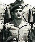 A formative black-and-white photograph of a young, confident-looking soldier. He wears a military-issue shirt and a dark-coloured beret, marked with insignia. He smiles into the camera.