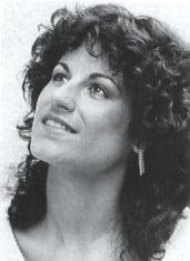 Three-quarter profile publicity shot of young woman with long, dark, curly hair, eye make-up and earrings, looking upwards, half-smiling.