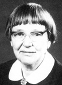 An older white woman with short straight hair and bangs; she is wearing eyeglasses, a white collar, and a small pin or pendant.