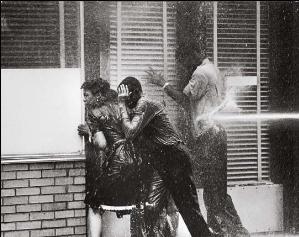Three black high school students, two boys and a girl, facing into a storefront window to avoid being hurt by a water cannon blasting a boy at his back; all three are dripping with water