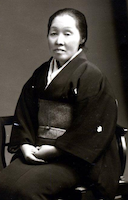 Jepang wanita setengah baya, mengenakan kimono gelap, duduk, dengan tangan terlipat di pangkuannya.