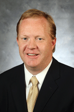 Man with dark blonde hair wearing a black suit jacket, white dress shirt, and a beige necktie