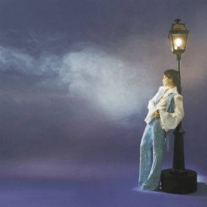 A short-haired woman wearing sequined trousers resting her back against a lit up lamppost.