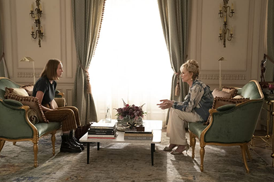 The screenshot is a wide shot of a lavish living room where Ava Daniels and Deborah Vance are seated opposite each other on identical green couches separated by a marble coffee table. The shot shows their profiles.