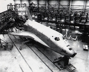 A black and white photo shows a Shuttle-shaped object with a more metalic presentation and missing its rounded nose cone resting in a warehouse surrounded on two sides by long and square metal scaffolding.