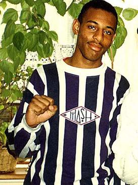 Photograph of a young Afro-Caribbean male, cropped to show his chest and head. He has black hair, shaved very short, and a slight moustache. He is wearing a navy-and-white vertically striped crew neck shirt. He is standing in front of a large indoor plant.