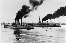 Zwei Schlachtschiffe dampfen in einen Hafen mit mehreren kleineren Booten um sie herum.  Das größere Schlachtschiff ist im Hintergrund, während das kleinere im Vordergrund steht.