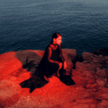 Kourtesis kneeling on rocks overlooking the sea with a red light shining on her