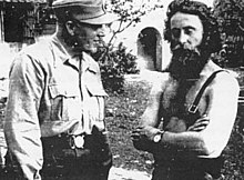 an SS officer in shirtsleeves and a mountain cap standing next to a bearded bare-chested man
