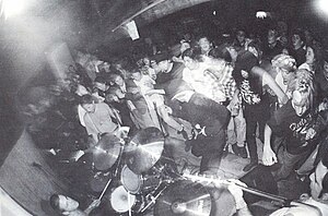 A Fishwife show from 1992 picturing Chris Prescott (on drums, far left), Ryan Foxe (with microphone, black shirt, and cap in front of drum set), Gar Wood (with guitar, left of drum set), and Matt Ohlin (with bass guitar, just visible in lower right corner). Photo by Mark Waters.